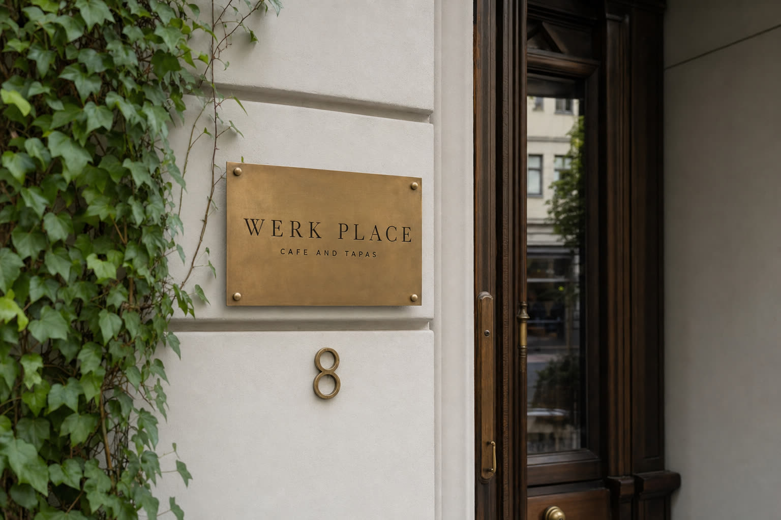 Wejście do Werk Place & Tapas: mosiężna tabliczka z grawerowanym napisem WERK PLACE CAFE AND TAPAS na białej ścianie obok ciemnych drewnianych drzwi, numer 8, gałązki bluszczu